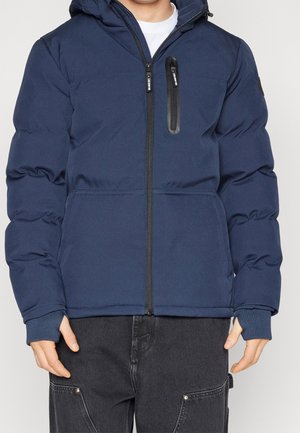 Homme portant une veste rembourrée zippée bleu marine avec une poche poitrine verticale et un jean foncé avec des poches avant visibles.