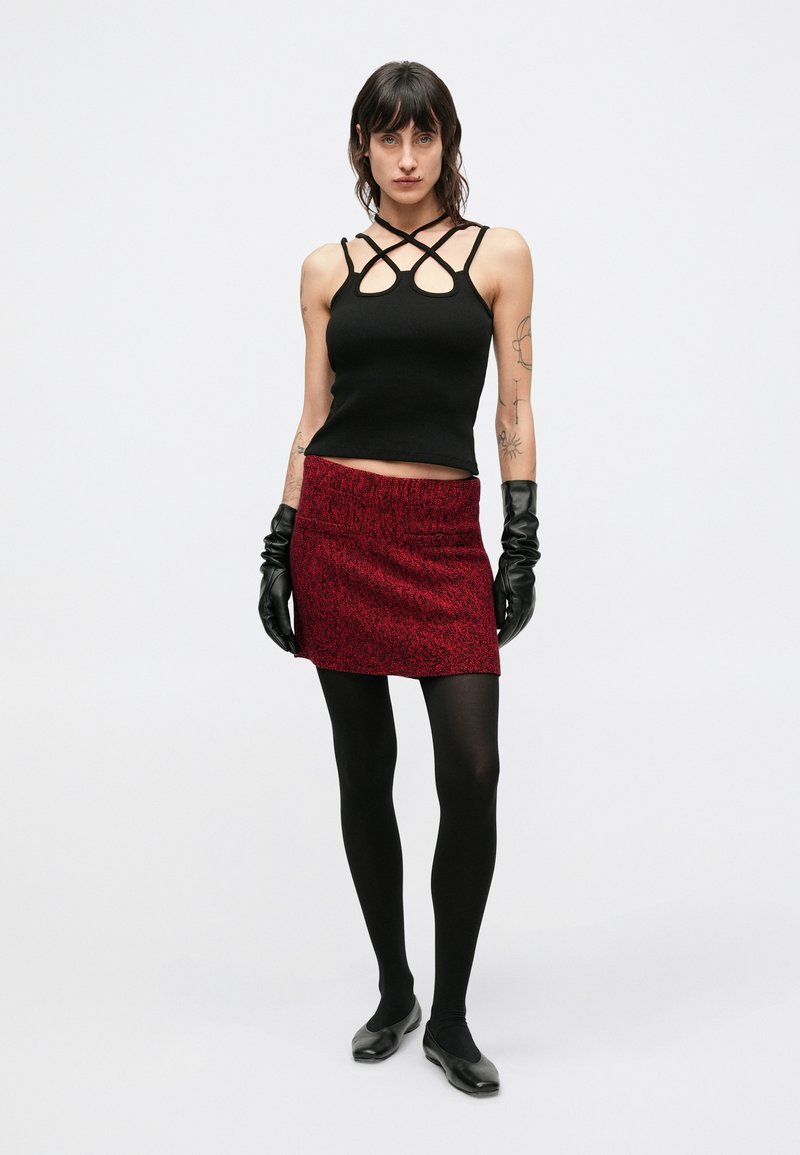 Black ribbed halter top with crisscross straps, paired with a short red skirt featuring a textured pattern, black tights, and gloves.