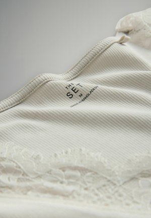 Krém színű bordázott anyag csipkével a tetején, rajta egy nyomtatott címke: "THE SET M, MADE IN BANGLADESH." Puha tapintású, ívelt nyakkivágással.