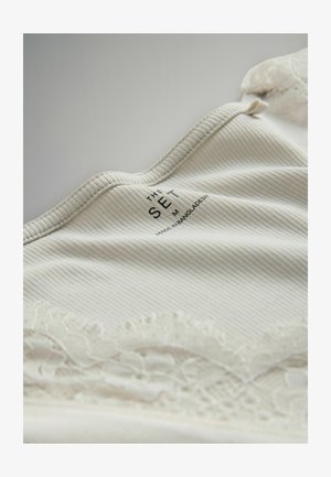 Krämfärgad ribbad tyg med spetskant upptill och en tryckt etikett med texten "THE SET M, MADE IN BANGLADESH." Mjuk textur, rundad halsringning.