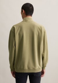 Man in een olijfgroen sweatshirt en zwarte jeans die met zijn rug naar de camera staat tegen een effen beige achtergrond.