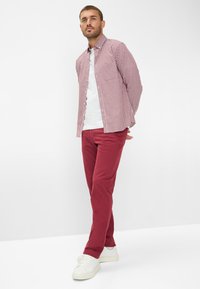 Camicia a quadretti rossa e bianca sopra una maglietta bianca, abbinata a pantaloni rossi e sneakers bianche. Il modello si trova in piedi con le mani dietro la schiena.