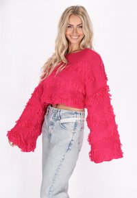 Fuchsias oversized cropped tröja med texturerad fransdetalj och vida ärmar, ihop med ljusblå högmidjade jeans.