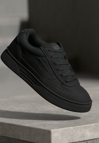 Zwarte sneaker met een bovenwerk van glad leer, ronde neus, platte vetersluiting en een gestructureerde buitenzool. Bevat minimaal merk en zijdetails.