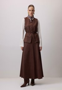 Rjava obleka iz blaga z oblečenim telovnikom z gumbi, poudarjenim pasom in dvema žepoma. Stilizirano z belo srajco in vzorčastim šalcem.