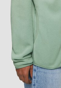 Hellgrüner Fleece-Jacke mit weichem Textur, Rundsaum und umgeschlagenen Ärmeln, kombiniert mit Jeans.