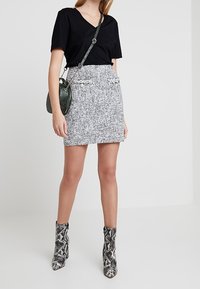 Černé tričko s v-dekoltem, přiléhavá sukně z šedého a bílého tkaného materiálu, kotníkové boty s hadím vzorem a zelená crossbody kabelka. Jednoduchý, strukturovaný vzhled.