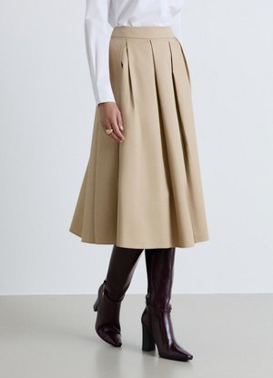 Gonna midi plissettata beige indossata con stivali marrone scuro con tacco fino al ginocchio e camicia bianca a maniche lunghe, mano appoggiata al fianco con anello d'oro.
