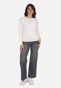 Maglione bianco a treccia con maniche lunghe abbinato a jeans a zampa d'elefante blu scuro e sneakers bianche, indossato da una modella femminile su uno sfondo neutro.