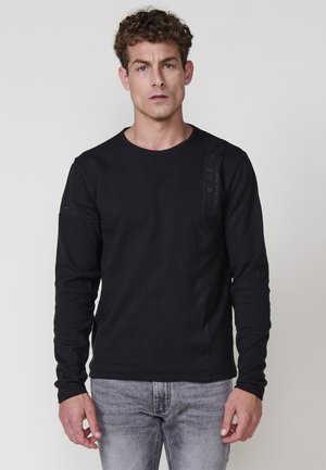 Camiseta negra de manga larga con escote redondo, textura de canalé y panel lateral que presenta gráficos sutiles. Combinada con jeans de mezclilla en gris claro.