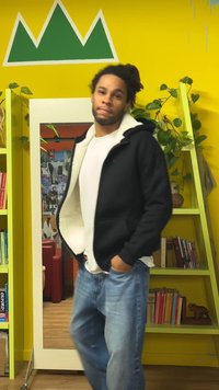 Schwarzer, mit Fleece gefütterter Kapuzenpullover mit weißem Innenfutter, getragen über einem weißen Hemd. Blaue Jeans. Grüne Wand mit Bergmotiv. Spiegel und Bücherregale.