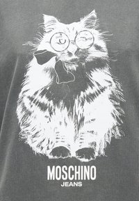 Αφράτη γάτα που φορά στρογγυλά γυαλιά και παπιγιόν, εικονογραφημένη με λευκό χρώμα πάνω σε γκρι ύφασμα με το κείμενο "MOSCHINO JEANS" από κάτω.