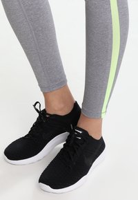 Piernas con leggings grises con una franja verde neón y zapatillas deportivas negras con suelas blancas sobre un fondo blanco.
