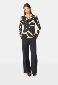 Maglietta a maniche lunghe color crema e nero con motivo floreale astratto; abbinata a pantaloni neri su misura. Il modello è in piedi con le mani nelle tasche.