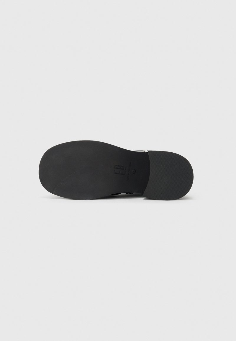 Semelle de chaussure noire avec une texture en caoutchouc lisse et un motif de relief minimal. Présente un design plat et une empreinte de logo discrète.