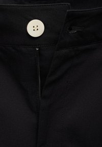 Pantaloni neri con zip e una vita ampia decorata da un bottone color crema. Tessuto liscio con una leggera trama.