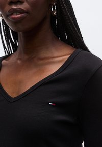 Musta, ribbineulottu pitkähiha-paita, jossa on V-pääntie ja pieni kirjailtu logo punaisessa, valkoisessa ja sinisessä kaula-aukon lähellä.