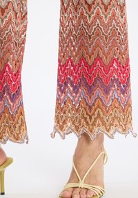 Kanten broek met zigzagpatroon in veelkleurige tinten van oranje, roze en paars; scalloped zoomdetail. Gecombineerd met gele sandalen met bandjes.