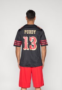 Μαύρη ποδοσφαιρική φανέλα με κόκκινες και χρυσές λεπτομέρειες, με το όνομα "PURDY" και τον αριθμό "13" στην πλάτη, συνοδευόμενη από κόκκινα σορτς.