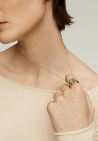 Collar de cadena de oro con un colgante de gema acuática cuadrada, usado con una blusa beige; un anillo de oro que presenta una piedra azul similar en la mano.