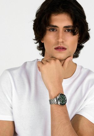 Orologio in acciaio inossidabile con quadrante verde, dettagli argentati e bracciale metallico. Indossato su un polso nudo contro uno sfondo di t-shirt bianca.