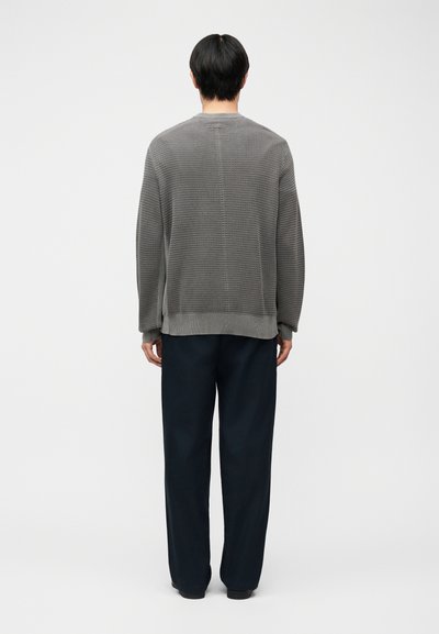 Homme aux cheveux noirs courts, de dos, portant un pull gris texturé et un pantalon noir ample, sur un fond blanc uni.