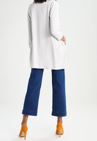 Long manteau gris clair à manches trois-quarts, associé à un jean large bleu foncé et des chaussures à talons hauts orange ; détail de poche visible.