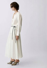 Chemise en lin blanche, légère, à manches longues et col, associée à une longue jupe blanche fluide et des chaussures à talons hauts à motifs.