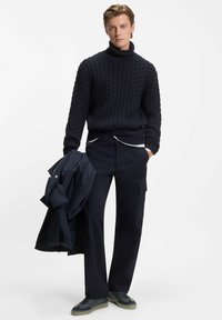 Marine gebreide coltrui met een textuurpatroon, gecombineerd met donkere pantalon en een lichtgewicht jack. Casual schoeisel met een grijze zool.