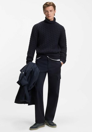 Marine gebreide coltrui met een textuurpatroon, gecombineerd met donkere pantalon en een lichtgewicht jack. Casual schoeisel met een grijze zool.