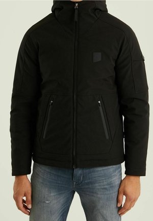 Czarna kurtka z kapturem, zapinana na zamek błyskawiczny z przodu, z dwoma kieszeniami na zamek, noszona przez osobę w niebieskich dżinsach, stojącą na tle jednolitego jasnego tła.