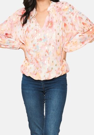 Mujer con una blusa floral rosa translúcida con detalles dorados y vaqueros azules, posando con las manos en las caderas sobre un fondo liso.