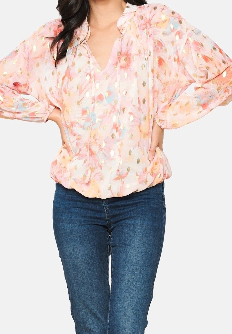 Femme portant un chemisier rose transparent à motif floral avec des accents dorés et un jean bleu, posant avec les mains sur les hanches devant un fond uni.