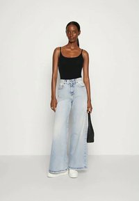 Kobieta stoi w czarnym topie na ramiączkach, jasnoniebieskich dżinsach z wysokim stanem i szerokimi nogawkami, białych sneakersach, trzymając małą czarną torbę.