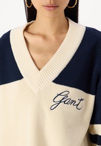 Krémový a námořnicky modrý pletený svetr s výstřihem do V, žebrovaným límcem a výraznými vodorovnými pruhy. Na hrudi má vyšitý nápis "Gant".