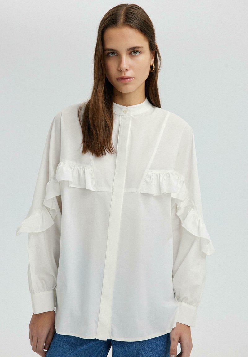 Camicia bianca con bottoni, con accenti arricciati sul petto e sulle maniche. Realizzata in materiale morbido, presenta un colletto alla mandarino e maniche lunghe.