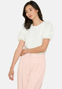 Balta T-kreka ar teksturētām, puffa piedurknēm ziedu rakstā, kombinēta ar gaiši rozā biksēm, kurām ir brīvs piegriezums un apaļa kakla izgriezuma forma.