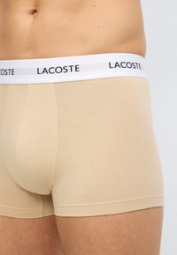 Ανδρικά μποξεράκια μπεζ με λευκή μέση που φέρει το μαύρο λογότυπο "LACOSTE", φορεμένα από έναν άνδρα με ορατό το κάτω μέρος του κορμού και το πάνω μέρος του μηρού.