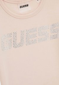 Vaaleanpunainen T-paita, joka on valmistettu pehmeästä kankaasta ja jossa etuosassa on suuri, kiiltäviä kiviä sisältävä "GUESS" -logo. Pyöreä pääntie ja lyhyet hihat.