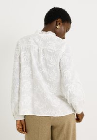 Blusa blanca con bordado floral texturizado, mangas largas y un collar con borde de encaje. La parte posterior presenta un canesú y un corte holgado.