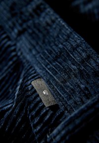 Donkerblauwe corduroystof met verticale ribbels, voorzien van een klein zwart label met een wit logo. Zachte textuur met opvallende patroon details.