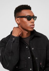 Giacca di jeans nera con collo in pelliccia; indossata sopra una camicia nera. La persona indossa occhiali da sole neri con dettagli laterali.
