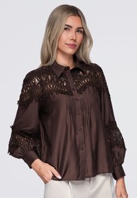 Femme aux longs cheveux blonds portant un chemisier brun foncé avec des détails en dentelle sur les épaules et les manches, debout avec une main dans la poche.