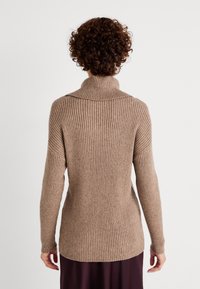 Une personne aux cheveux bouclés vue de dos, portant un pull en maille côtelée beige à manches longues avec un col roulé et un pantalon sombre, sur fond uni.