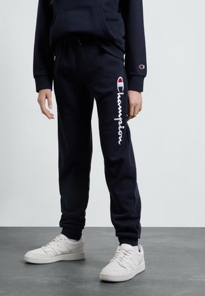 Osoba ubrana w czarne spodnie dresowe Champion i białe buty sportowe New Balance, stojąca na szarej podłodze na tle jednolitego jasnego tła.