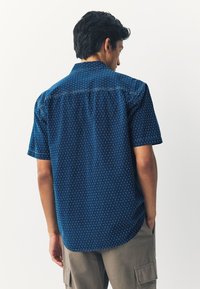 Chemise à manches courtes en tissu bleu foncé avec un motif blanc répétitif. Col tailleur et ourlet courbé ; détail de pli au dos.