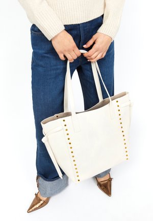 Borsa a spalla bianca in pelle con borchie dorate lungo i lati, doppio manico e design aperto in cima. Indossata con jeans blu scuro e scarpe marroni.