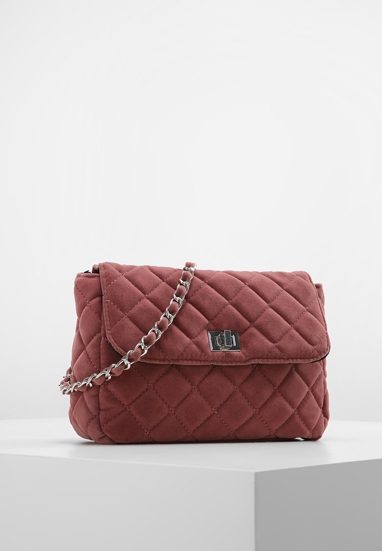 Borse in tessuto rosa polveroso trapuntata con tracolla in catena argentata e chiusura rettangolare in metallo, esposta su un piedistallo bianco contro uno sfondo semplice.