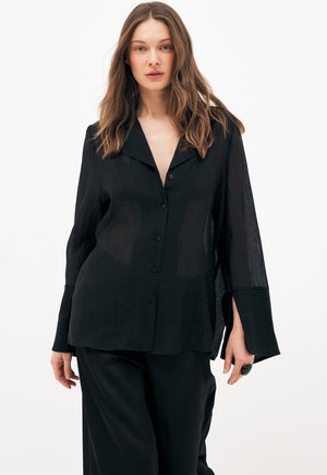 Femme aux cheveux ondulés portant une chemise noire ample et transparente à boutons et un pantalon noir, debout devant un fond clair.