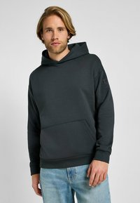 Sudadera con capucha de color gris oscuro, con un bolsillo frontal, puños acanalados y un acabado texturizado. Presenta un logotipo grabado en la manga.
